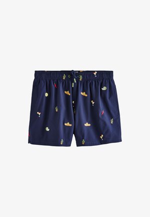 Man draagt blauwe zwemshorts met geborduurde cocktails, chilipepers, cactussen en sombrero's, staand op een zanderig strand.