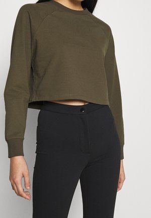 Kvinde iført en olivengrøn cropped sweatshirt med lange ærmer og højttaljede sorte bukser, stående mod en ensfarvet lys baggrund.