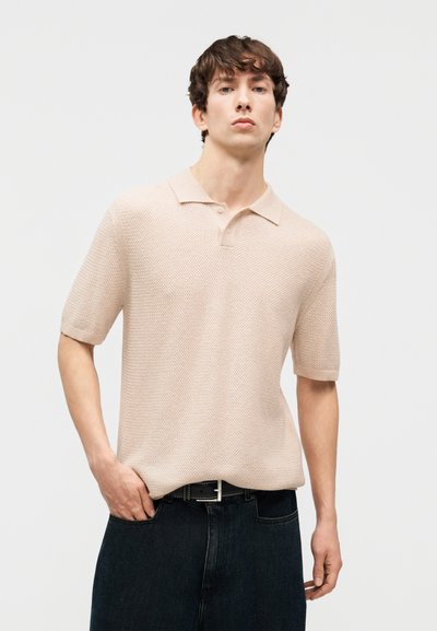 Νεαρός άνδρας που φοράει μπεζ πόλο με κοντά μανίκια και υφή, μαύρη ζώνη και σκούρο τζιν, στέκεται μπροστά σε ένα απλό φωτεινό φόντο.