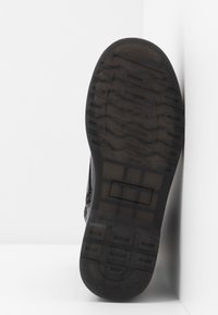 Semelle d'une botte noire avec un motif de bande de roulement texturé, légèrement inclinée contre un fond blanc d'angle.