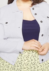 Vit denimjacka med svarta knappar, utrustad med två bröstfickor. Bärs över en mörkblå topp, ihop med en gul kjol med blommönster.