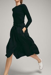 Femme portant un haut noir à manches longues, une jupe noire taille haute avec poches et des baskets blanches, photographiée en plein mouvement sur un fond uni.