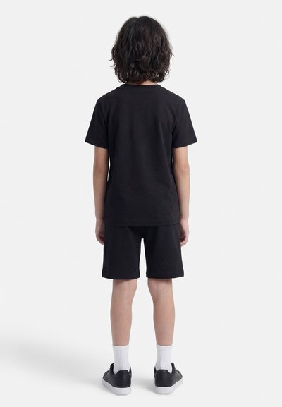 Παιδί με μεσαίου μήκους σκούρα μαλλιά στέκεται γυρισμένο από πίσω, φοράει μαύρο μπλουζάκι, μαύρο σορτς, λευκές κάλτσες και μαύρα αθλητικά παπούτσια σε λευκό φόντο.