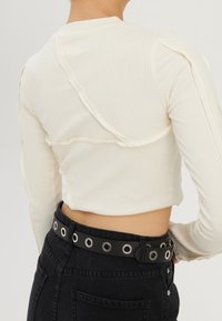 Rückansicht einer Person, die ein cremefarbenes, geripptes Langarm-Crop-Top und hochgeschnittene schwarze Jeans mit einem schwarzen Gürtel mit großen silbernen Ösen trägt.