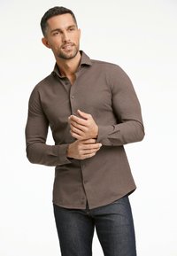 Camisa marrón de botón con ajuste slim, mangas largas y una textura sutil, combinada con jeans de mezclilla oscuros, presentada sobre un fondo neutro.