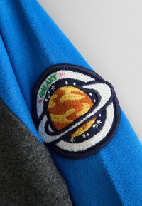 Farebná modrá a šedá tkanina s kruhovou vyšívanou nášivkou zobrazujúcou planétu s prsteňmi a hviezdami, zdôrazňujúca tému vesmíru.