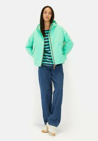 Licht blauwe gewatteerde jas met capuchon, met een ritsluiting en grote zakken, gecombineerd met een gestreepte marineblauwe en groene shirt en blauwe wijde jeans.