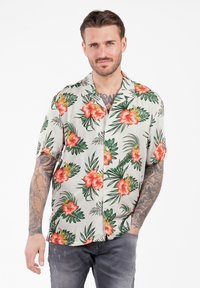Mann mit Tattoos, der ein kurzärmliges beiges Hawaiian-Shirt mit roten Hibiskusblumen und grünen Blättern trägt, die Hände in den Taschen, vor einem weißen Hintergrund.