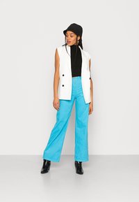 Blazer branco sem mangas com botões pretos, combinado com uma blusa preta canelada e calças de veludo cotelê largas em azul claro. Botas de tornozelo pretas.