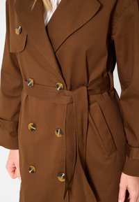 Cappotto trench marrone in cotone, con design doppiopetto, grandi bottoni, cintura e tasche laterali. Texture liscia e colletto classico.