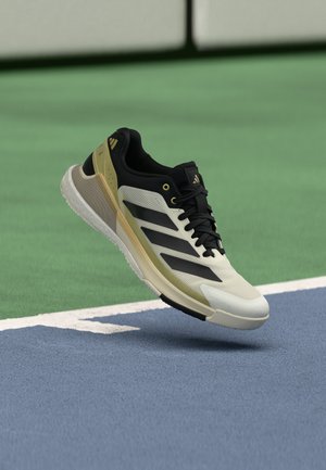Un único zapato deportivo ligero en negro, blanco y dorado con las rayas de Adidas parece flotar sobre una superficie de pista de tenis azul y verde.