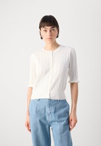 Cardigan bianco a maniche corte con texture a coste, scollatura ondulata e chiusura con bottoni. Le dettagli in pizzo lungo le maniche aggiungono interesse visivo.