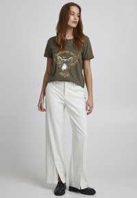 Olivengrøn t-shirt med ørn grafik, parret med hvide vidde bukser med frontslids. Modellen har sorte sko på.