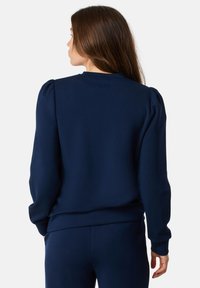 Donna con lunghi capelli castani, vestita con un felpa blu navy e pantaloni coordinati, vista di spalle su uno sfondo bianco uniforme.