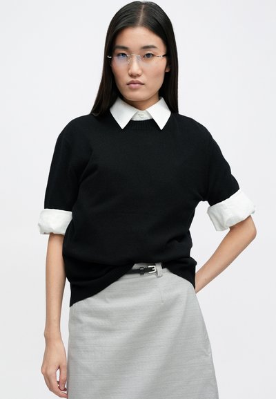 Μαύρο πλεκτό πουλόβερ με γυρισμένα μανίκια πάνω από ένα λευκό πουκάμισο. Το σύνολο ολοκληρώνεται με μια γκρίζα φούστα και μια γκρίζα ζώνη.