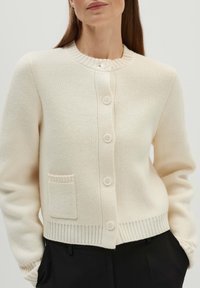 Cremfarbener Cardigan aus weichem Stoff, mit rundem Ausschnitt, fünf Knöpfen, geripptem Saum und einer kleinen Tasche auf der linken Seite.