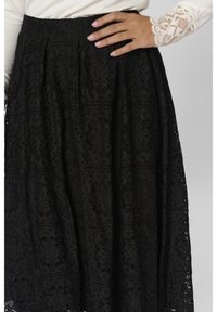 Zwarte kantrok met bloemmotieven, textuurstof en een tailleband. Een hand ligt op de rok, wat het gedetailleerde ontwerp benadrukt.