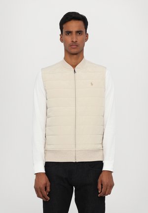 Gilet matelassé beige avec un zip sur le devant, un ourlet côtelé et un col rond. Présente un petit logo sur la poitrine. Porté par-dessus une chemise blanche à manches longues.