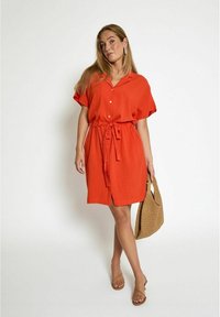 Bright orange shirt dress with a button-down front, short sleeves, tied waist, and lightweight fabric. Holds a textured brown woven bag.