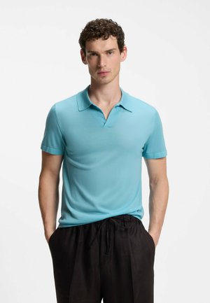 Homme aux cheveux courts et bouclés, portant un polo bleu clair à manches courtes et un pantalon noir à cordon, debout les mains dans les poches.