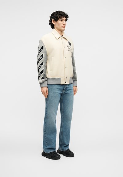 Νεαρός άνδρας με σγουρά μαλλιά, ντυμένος με ένα βελούδινο μπουφάν varsity σε εκρού και γκρι, με μαύρο κείμενο στα μανίκια και το στήθος, μπλε τζιν και μαύρα αθλητικά παπούτσια.