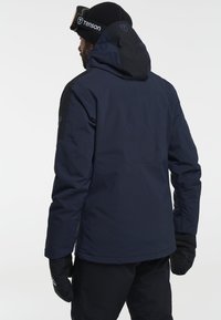 Man die met zijn rug naar de camera staat, gekleed in een marineblauwe en zwarte hoodie, een zwarte beanie met een bril, zwarte handschoenen en zwarte broek tegen een witte achtergrond.