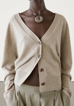 Dark-skinned person wearing a beige buttoned cardigan, matching pleated pants, and a round wooden pendant on a black cord.