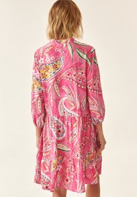 Vrouw met schouderlang blond haar, in een roze jurk met bloemen- en paisleypatroon en 3/4 mouwen, gezien vanuit de achterkant.
