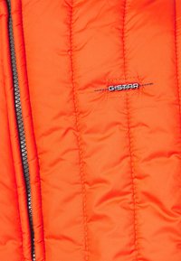 Jasně oranžová prošívaná bunda s viditelným vertikálním šitím, černým zipem a malou černo-bílou nášivkou s logem G-STAR.