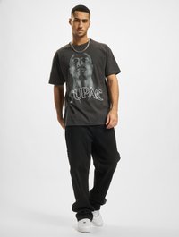 Grå bomulls t-skjorte med et stort hvitt grafikk av Tupac og teksten "TUPAC" nedenfor; sammen med løse svarte bukser og hvite sneakers.