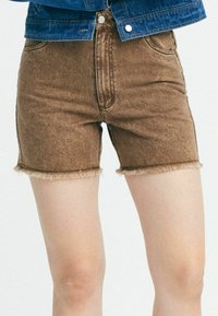 Pantalones cortos de mezclilla marrones con un dobladillo deshilachado, que presentan un diseño tradicional de cinco bolsillos y costura vertical en la parte frontal.
