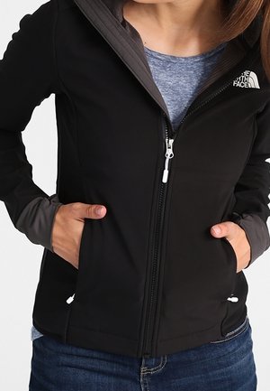 Henkilö pukeutuneena mustaan vetoketjulliseen North Face -takkiin sinisen paidan päällä, kädet takin taskuissa, nähtynä rinnasta lantioihin.