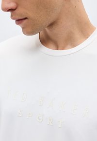 Κοντινό πλάνο στο κάτω μέρος του προσώπου και τον λαιμό ενός άνδρα που φοράει λευκό μπλουζάκι με στρογγυλή λαιμόκοψη και ανάγλυφο κείμενο "TED BAKER SPORT" στο στήθος.