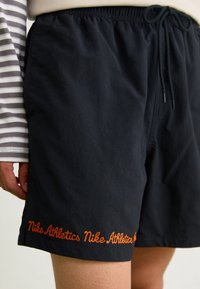 Person som bär svarta Nike Athletics-shorts med en orange broderad logotyp nära fållen och en randig långärmad tröja, beskuren vy.