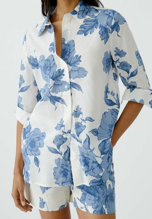 Woman wearing white button-up shirt and shorts with blue floral print, sleeves rolled to elbows, hands relaxed at sides.