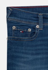 Tumma siniset farkut, joissa näkyvä ommel ja hopeiset metalliosat. Etutaskussa on punainen, valkoinen ja sininen logoväritys.