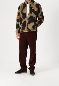 Man med fleecejacka i camouflage-mönster, vit skjorta, brun corduroybyxor och svarta sneakers som står mot en vit bakgrund.
