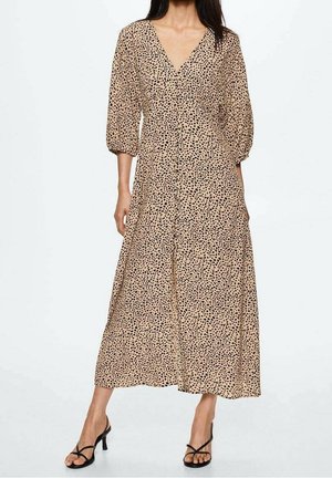 Vrouw draagt een beige midijurk met zwarte polkadots, driekwart pofmouwen, V-halslijn en zwarte sandalen met hak en bandjes.