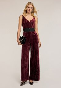 Burgundské sametové overal s širokým střihem, hlubokým výstřihem do V a černým opaskem. V kombinaci s malou černou kabelkou a špičatými botami.