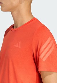 Camiseta deportiva roja con cuello redondo, textura suave, con un logo tonal en el pecho y tres rayas en el hombro.