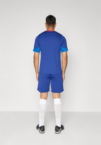 Blå trøye med korte ermer og rød halskant, kombinert med blå shorts. Hvite sokker og svarte treningssko fullfører antrekket.