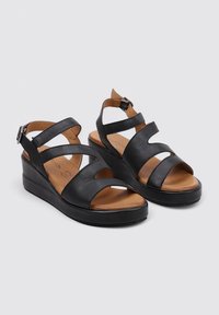 Sandalias de cuña de cuero negro con múltiples correas entrelazadas y cierre de hebilla. La plantilla es de color marrón claro con una textura suave.