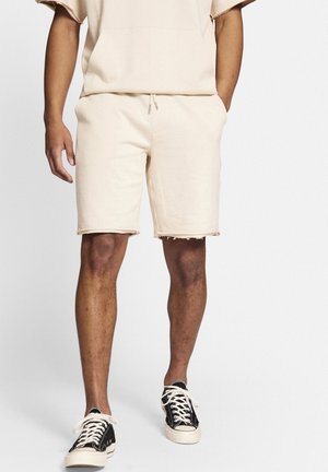 Mann trägt beige Kordelzug-Shorts und passendes Hemd mit schwarzen und weißen Schnürschuhen, steht vor weißem Hintergrund.