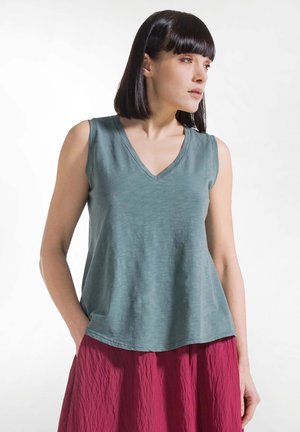 Donna con capelli scuri lisci che indossa una canotta smanicata verde acqua con scollo a V e una gonna rossa stropicciata, in piedi davanti a uno sfondo bianco.