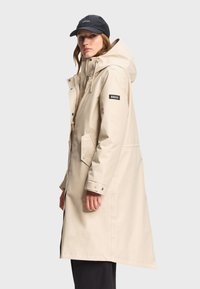 Femme portant un long imperméable beige à capuche avec poches avant et une casquette noire Aigle, debout devant un fond gris clair.