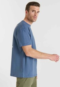 Hombre con camiseta azul y pantalones oliva extendiendo el brazo derecho, mirando ligeramente por encima del hombro contra un fondo claro y sencillo.