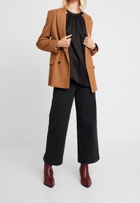 Blazer marrone su misura sopra una blusa nera a fantasia, abbinato a pantaloni neri a gamba larga e stivaletti bordeaux. Texture morbide e design strutturato.