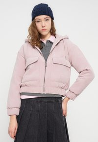 Ροζ μπουφάν από κορντόνι κομμένο στο ύψος της μέσης, με κουκούλα και τσέπες μπροστά, φορεμένο πάνω από γκρι πόλο και μαύρη πλισέ φούστα. Μπλε πλεκτός σκούφος στο κεφάλι.