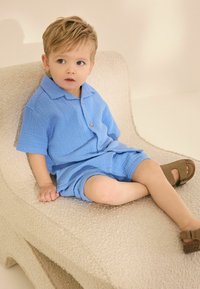 Camisa azul claro de manga corta y pantalones cortos a juego, hechos de una tela texturada. Sandalias marrones con hebillas. Sentado en una silla beige texturada.