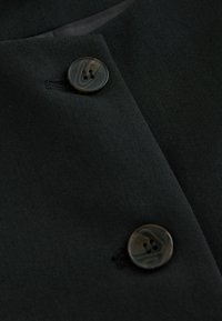 Lähikuva mustasta kankaasta, jossa on kaksi pyöreää nappia marmorikuviolla ja integroidut nappiaukot, sileä tekstuuri.